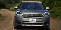 Front view of the Mini Cooper Electric Convertible driving on a dirt road surrounded by lush green hills, showcasing its modern design and electric capabilities.