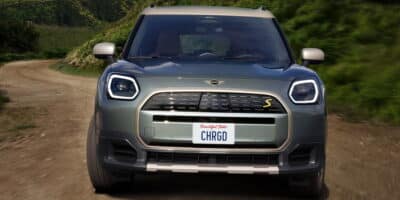 Front view of the Mini Cooper Electric Convertible driving on a dirt road surrounded by lush green hills, showcasing its modern design and electric capabilities.