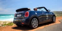 A 2024 Mini Cooper Convertible is parked on a coastal highway with red dirt and turquoise water.