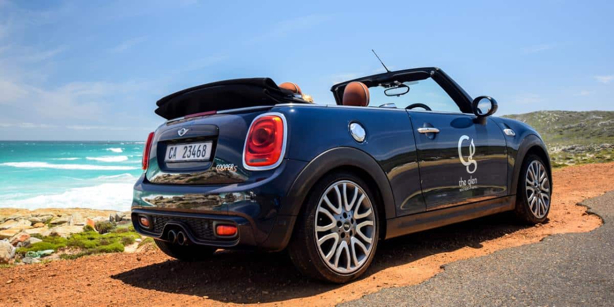 A 2024 Mini Cooper Convertible is parked on a coastal highway with red dirt and turquoise water.
