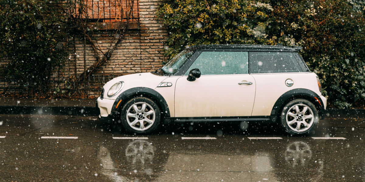 Side View Of beige white Color Mini Cooper MPG Parked On Street near old house. Snowy weather, snowfall.