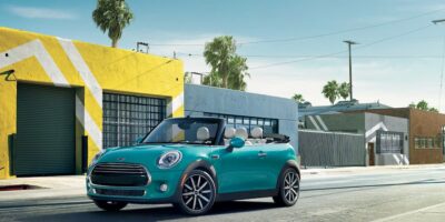 A turquoise 2025 Mini Cooper Convertible parked on a sunny street with colorful urban buildings and palm trees in the background.