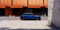 A sleek, blue 2025 Mini Cooper S parked on a quiet street in front of a modern orange building. The car's bold design and compact structure are emphasized in this urban setting, highlighting its sporty appeal