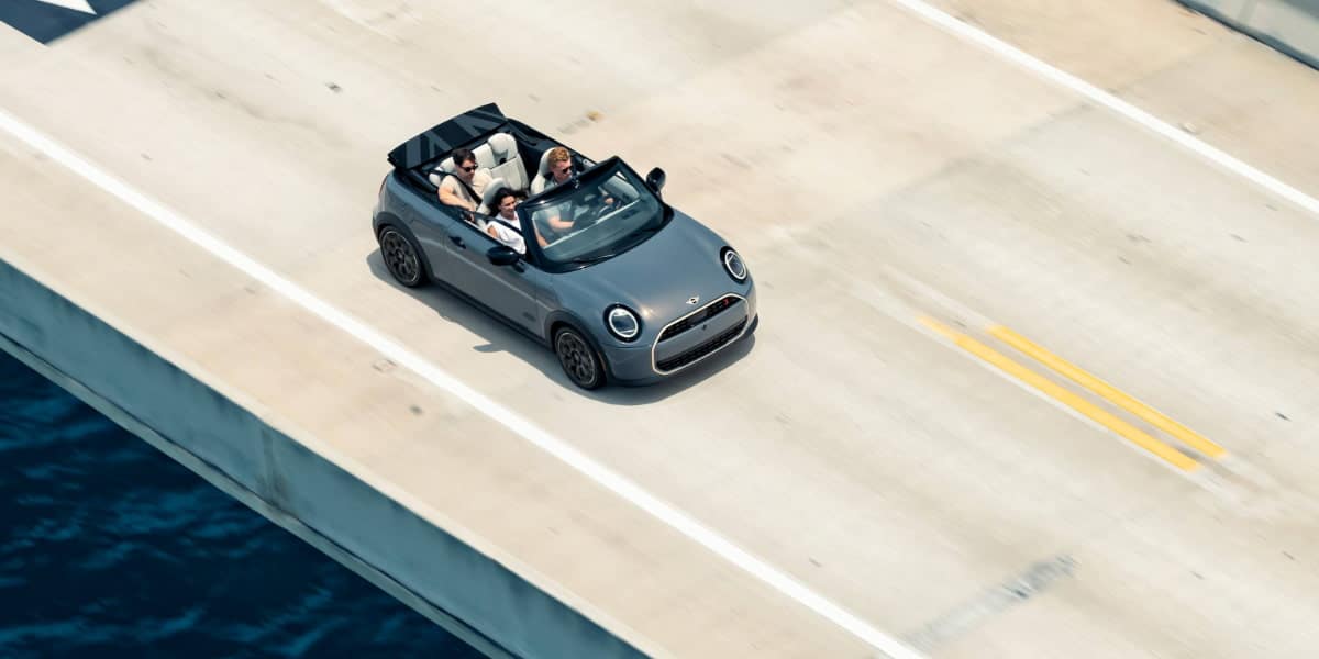 A MINI Cooper Convertible driving on a coastal highway with three passengers enjoying a scenic top-down ride. Though there is no 4 door MINI Cooper Convertible, this model offers an exciting alternative.