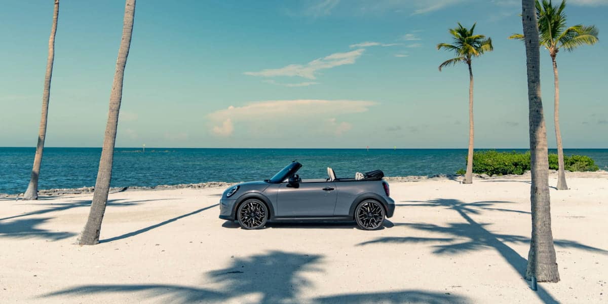 A gray MINI Cooper Convertible parked on a sandy beach with the ocean in the background. A 4 door MINI Cooper Convertible isn’t available, but this two-door model captures the open-air driving experience.