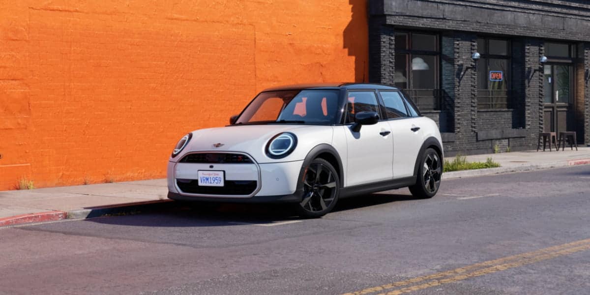 A modern white Mini Cooper parked on a city street with an orange wall in the background. The latest Mini Cooper generations showcase sleek design and advanced technology.