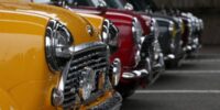 Row of brightly colored classic cars, featuring the original Mini Cooper at the front, parked outside in a drizzle at a Mini factory event