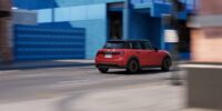 Rear three quarter view of a red 2026 Mini Cooper driving through a city street with modern buildings in the background