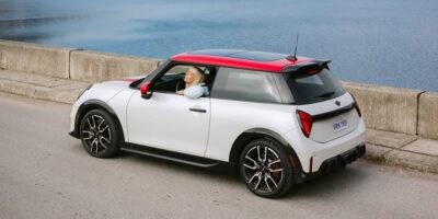 White two door hatchback driving along a waterfront road highlighting the sporty design of the 2026 Mini JCW with contrasting roof and wheels